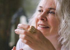 A senior woman with white hair holds a coffee cup, smiling thoughtfully indoors.